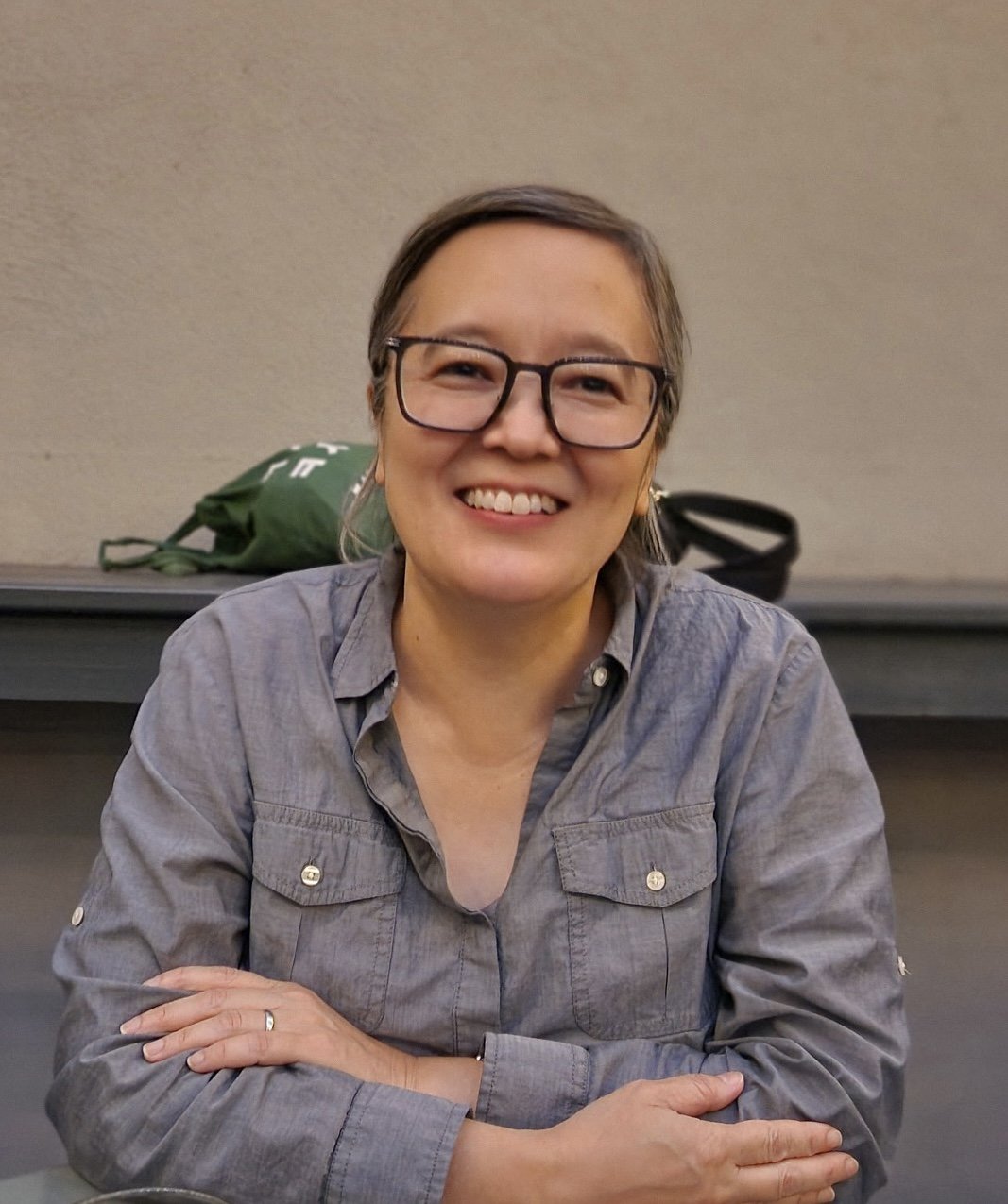 A woman with glasses smiling at the camera, sitting at a table with her arms crossed, in front of a beige wall.