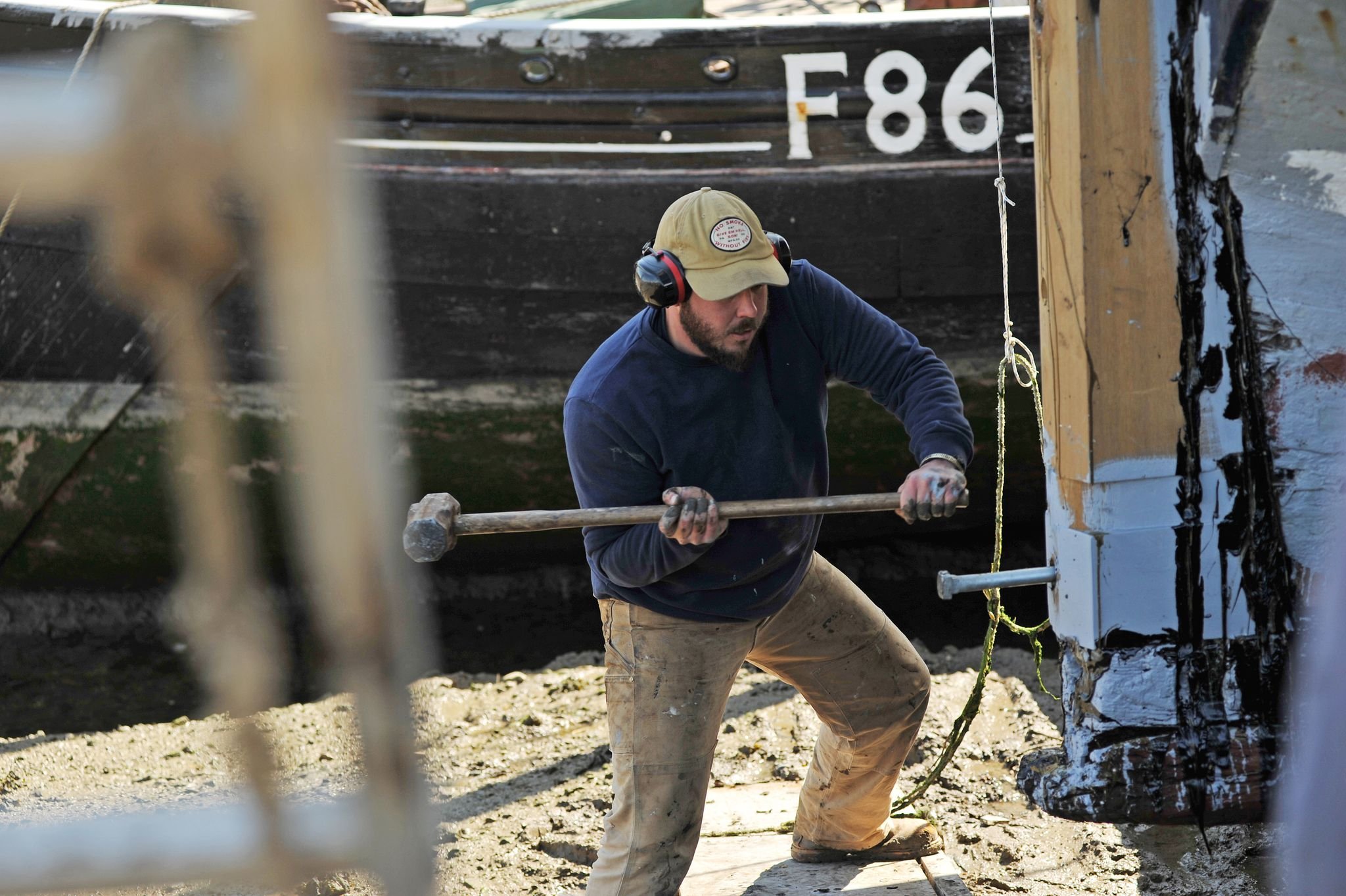 Lyndon March wearing a beige cap and hearing protection works outdoors with a sledgehammer next to a large black boat with the number F86 on it. The area appears to be a dock or shipyard, he is repairing the stern post of Thames Barge Edith May