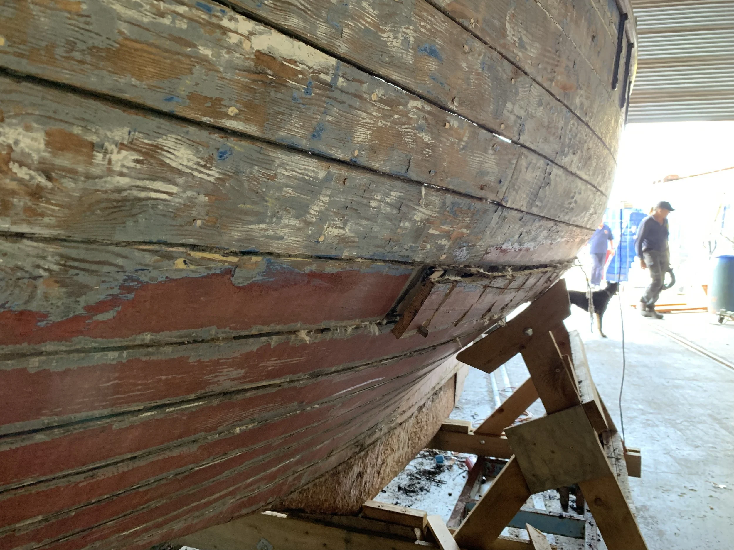 Close-up of weathered wooden boat hull with peeling red and blue paint on a boatlift, with people and equipment in the background at a boatyard.