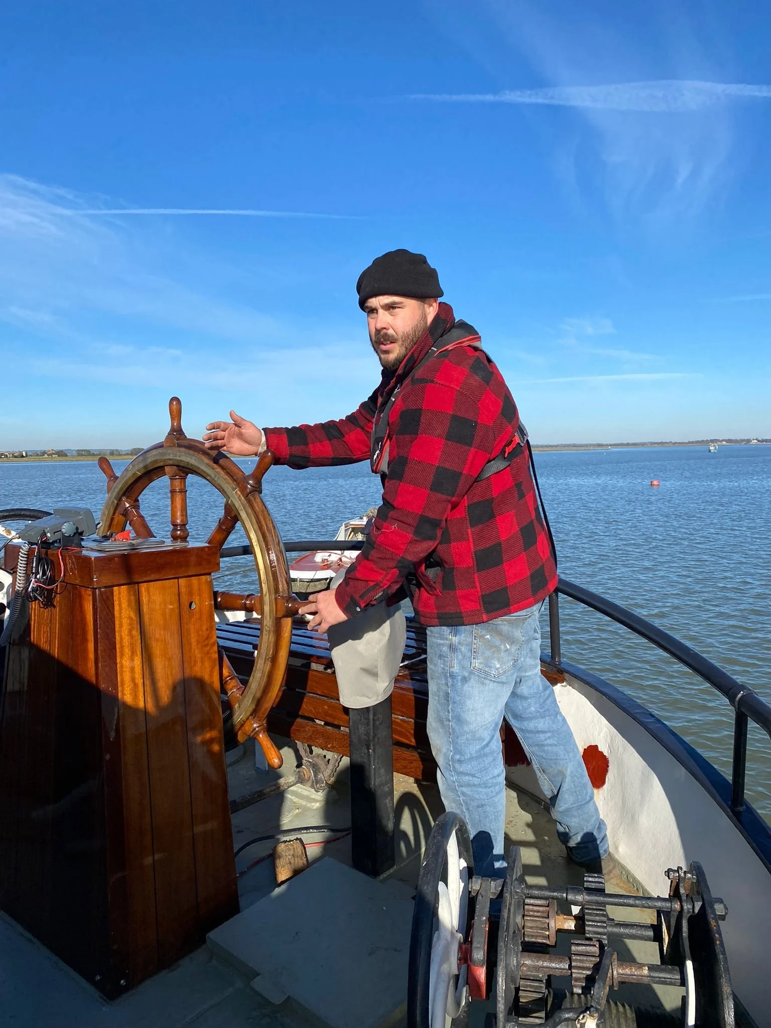 A man wearing a black beanie and red plaid jacket steering a boat by a large wooden ship's wheel, with water and a clear blue sky in the background.