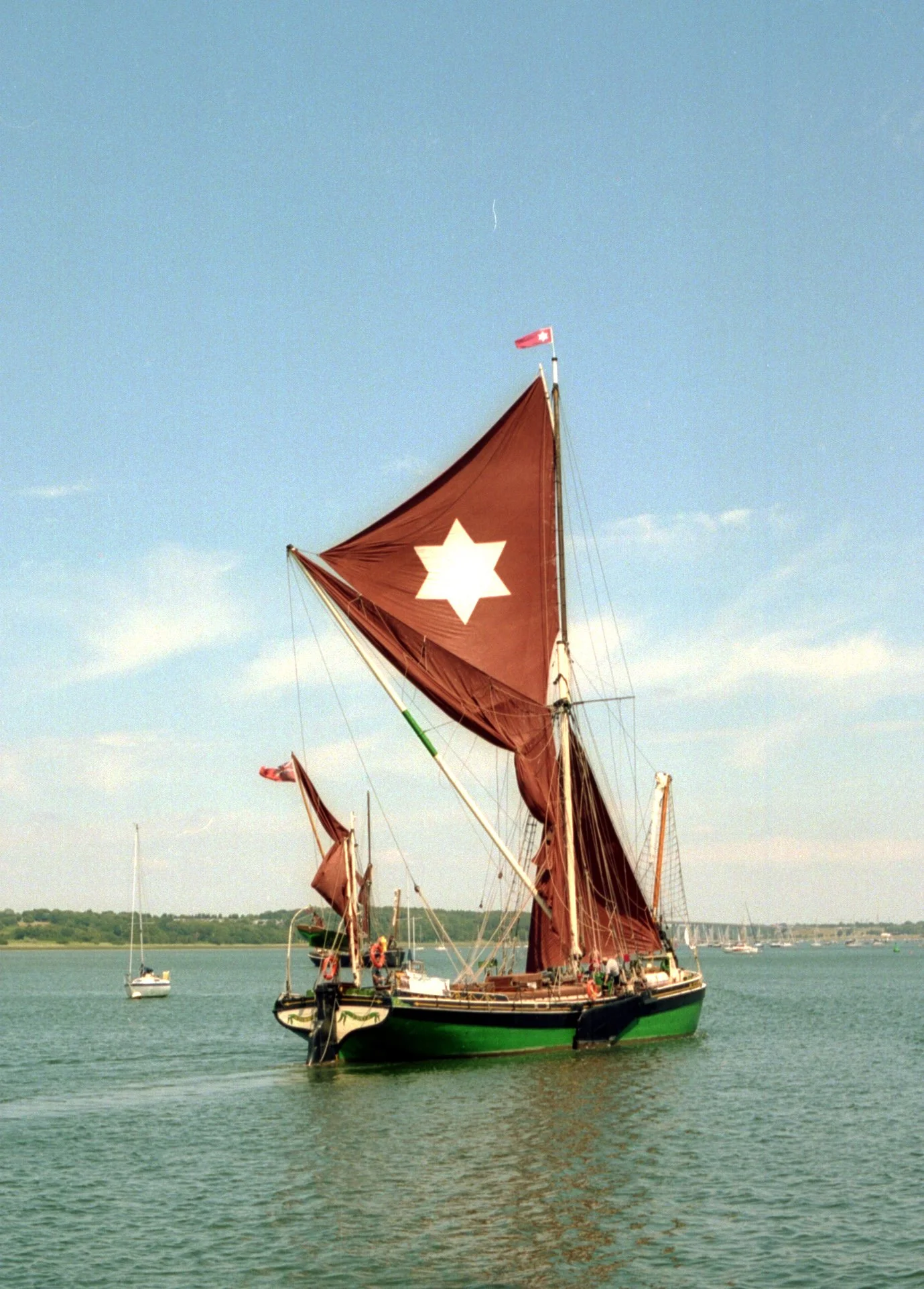 A traditional Thames Sailing Barge "The Kitty" with brown sails and a white star emblem, sailing on calm water with other boats and a tree-lined shoreline in the background.