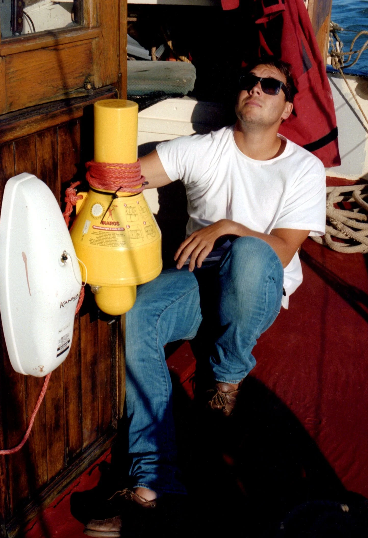 Lyndon Marchsitting on a boat deck, inside the wheelhouse of a Thames barge, wearing sunglasses and a white t-shirt, next to a yellow and red life preserver. Onboard Thames Sailing Barge Thistle