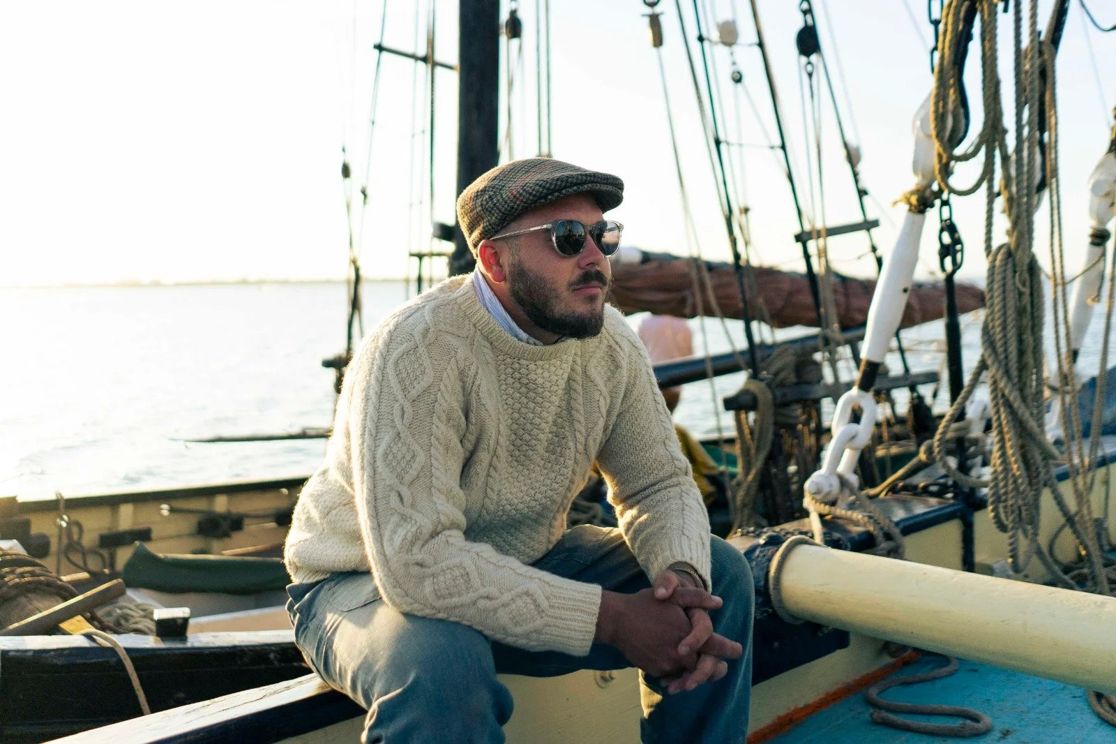 A man with a beard, wearing a checkered flat cap, round sunglasses, a cream cable-knit sweater, and gray pants, sitting on the edge of a boat with his hands clasped, near a body of water and sailboat rigging.