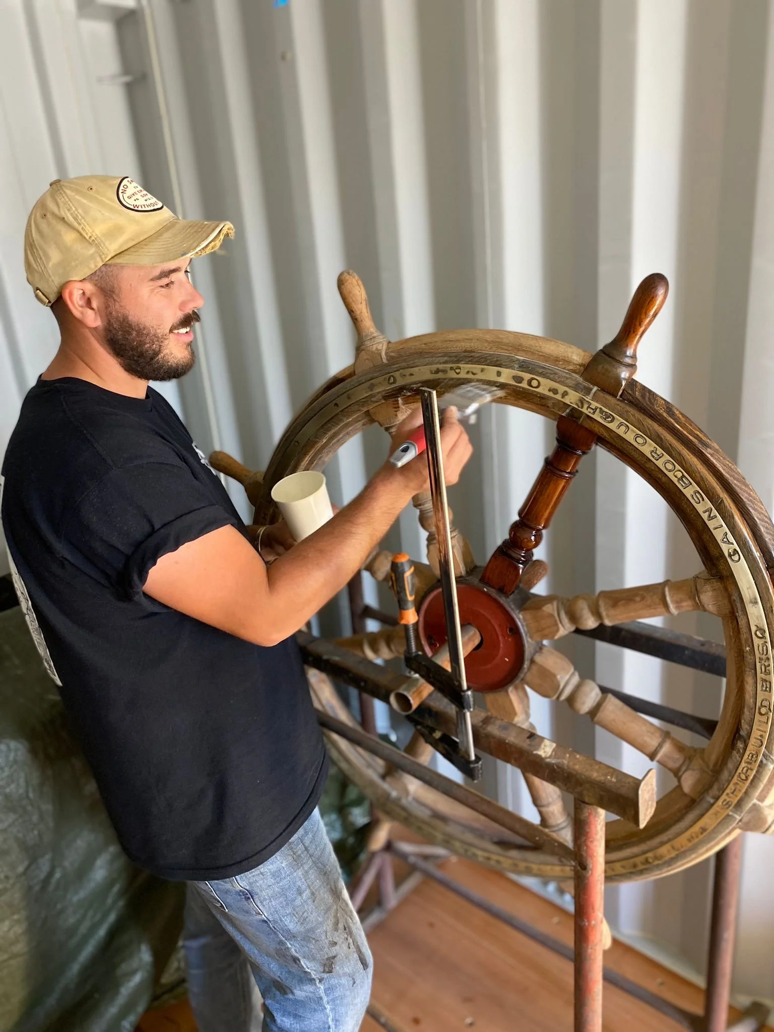 A man with a beige cap and black T-shirt standing in front of a large wooden ship wheel, using a marker to write or mark on the wheel.