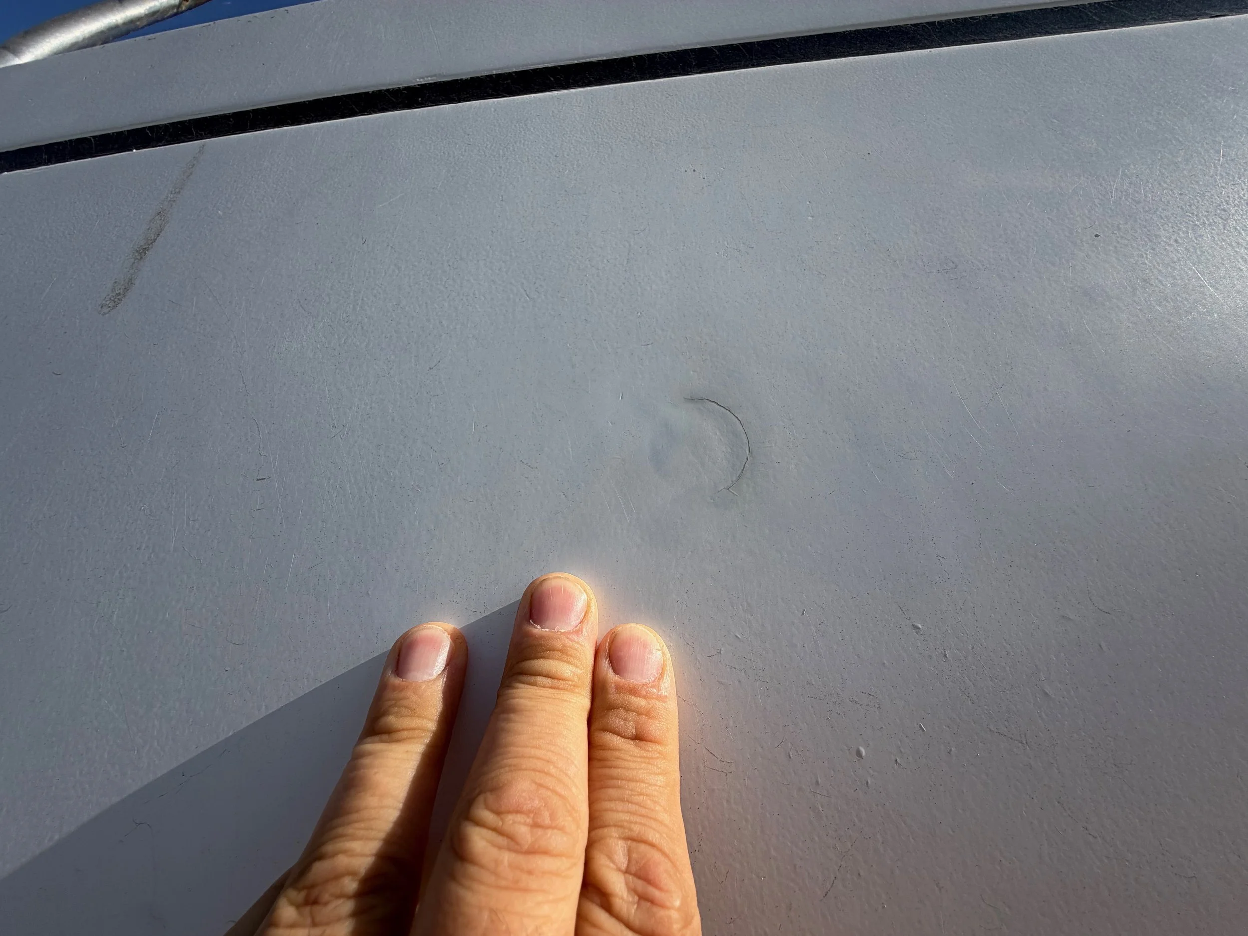 A person's hand with short fingernails holding a thin, gray, slightly curved wire or string against a light gray surface. The surface has some scratches and a small, faint hair or fiber.
