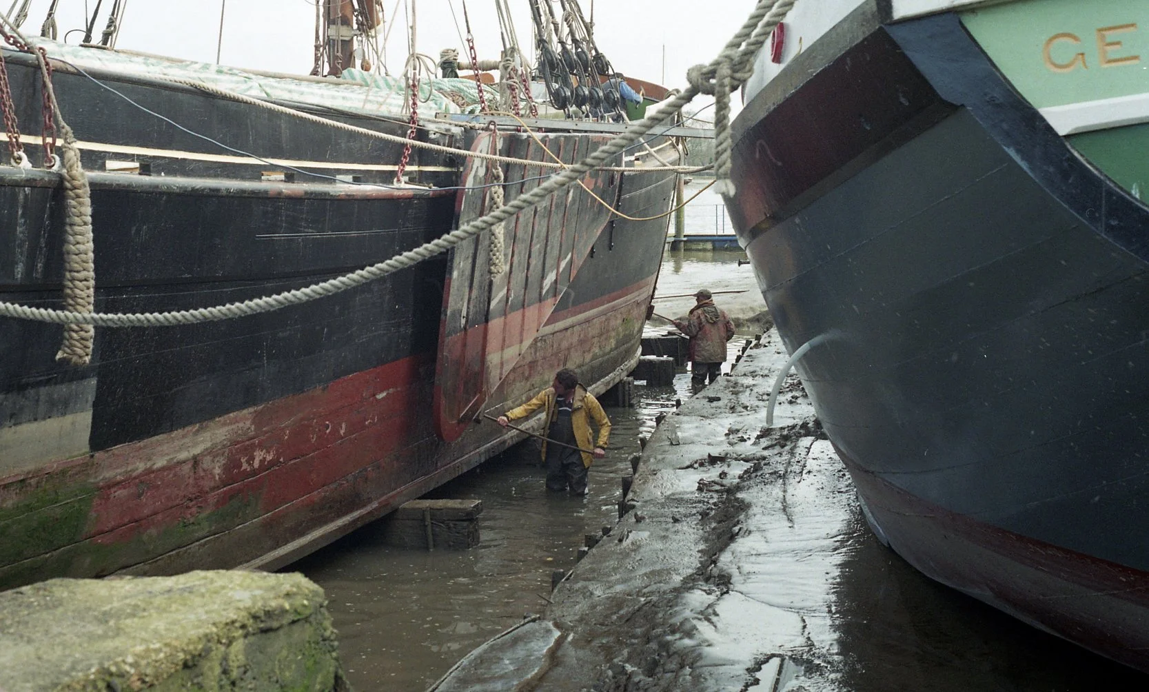 People working on boats in a muddy marina, cleaning and maintaining the ships docked at the harbor.