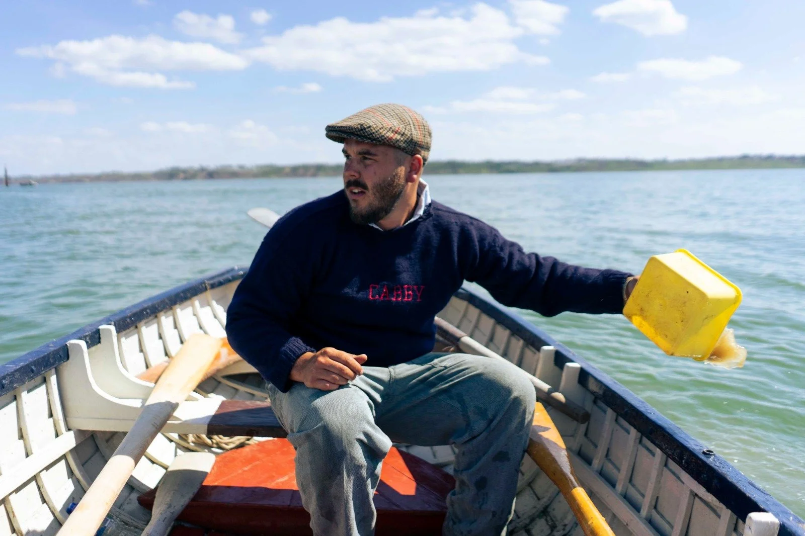Man sitting in a small boat holding a yellow bucket over the water on a sunny day, with a shoreline in the background.