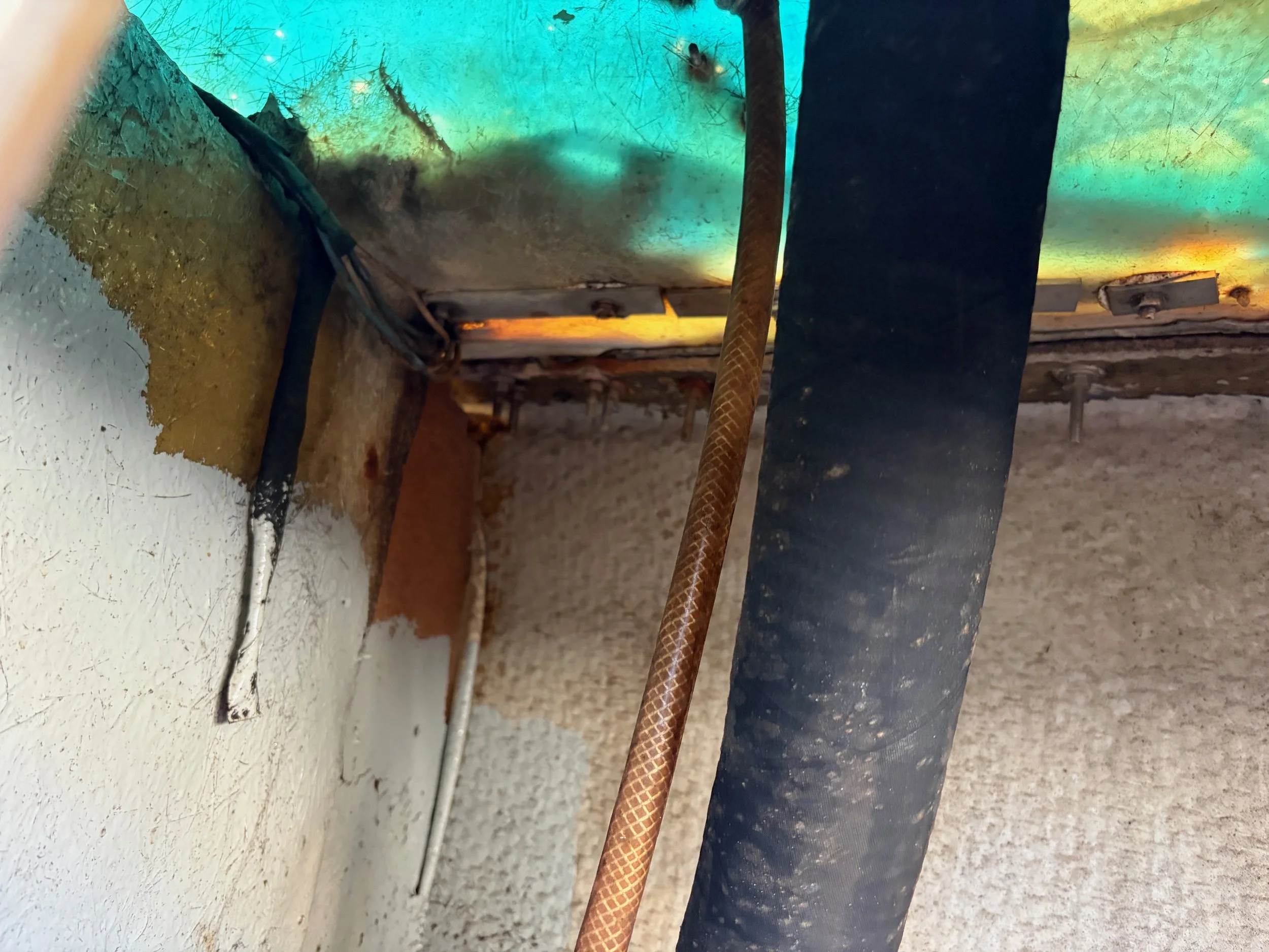 Close-up of electrical wires and cables coming out of the wall with a rusted pipe and black conduit pipe on a textured wall, with some colored lighting reflecting on the wall.