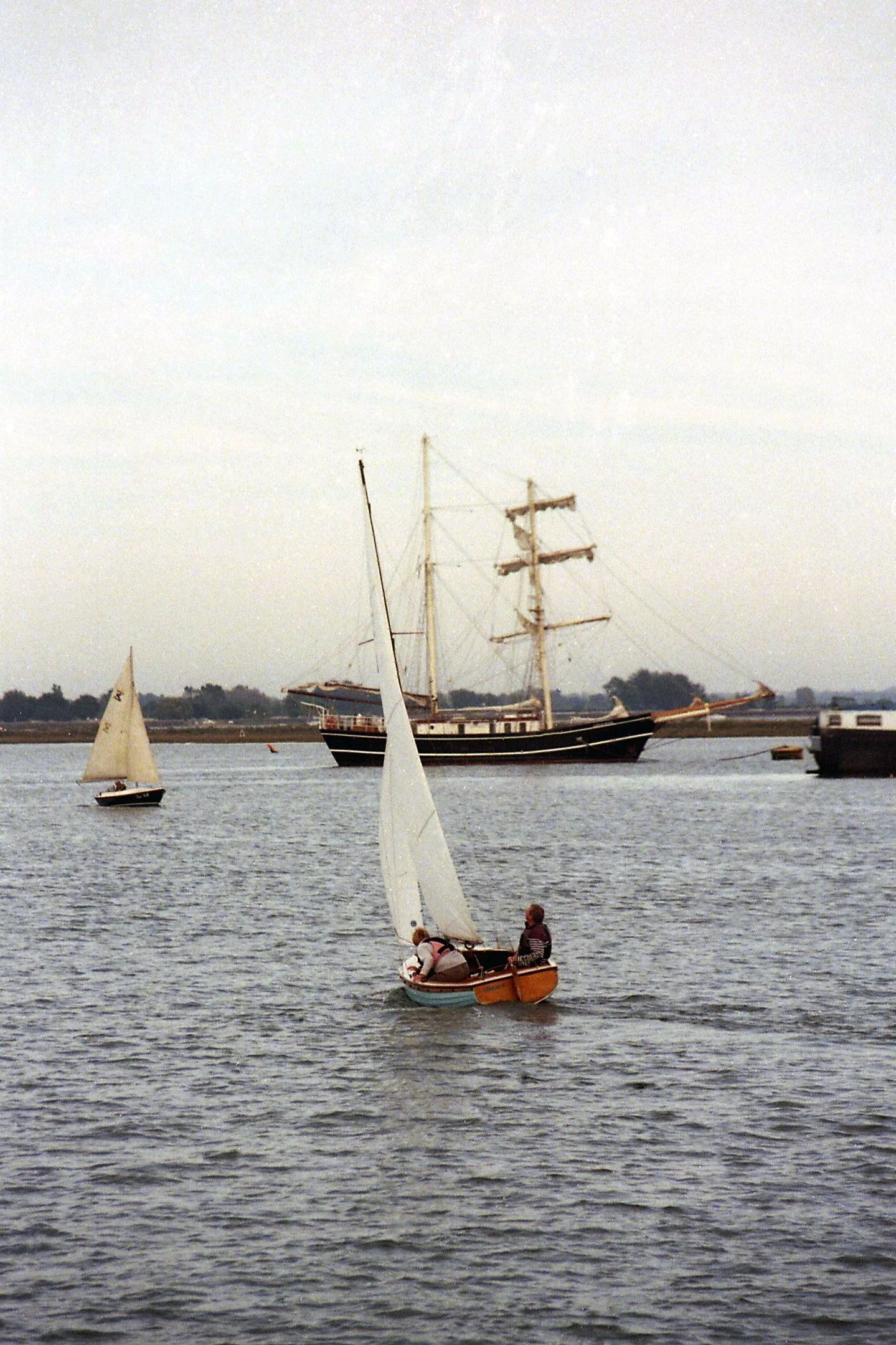 A sailboat with two passengers sailing in a body of water, with multiple sailing ships and a shoreline in the background.