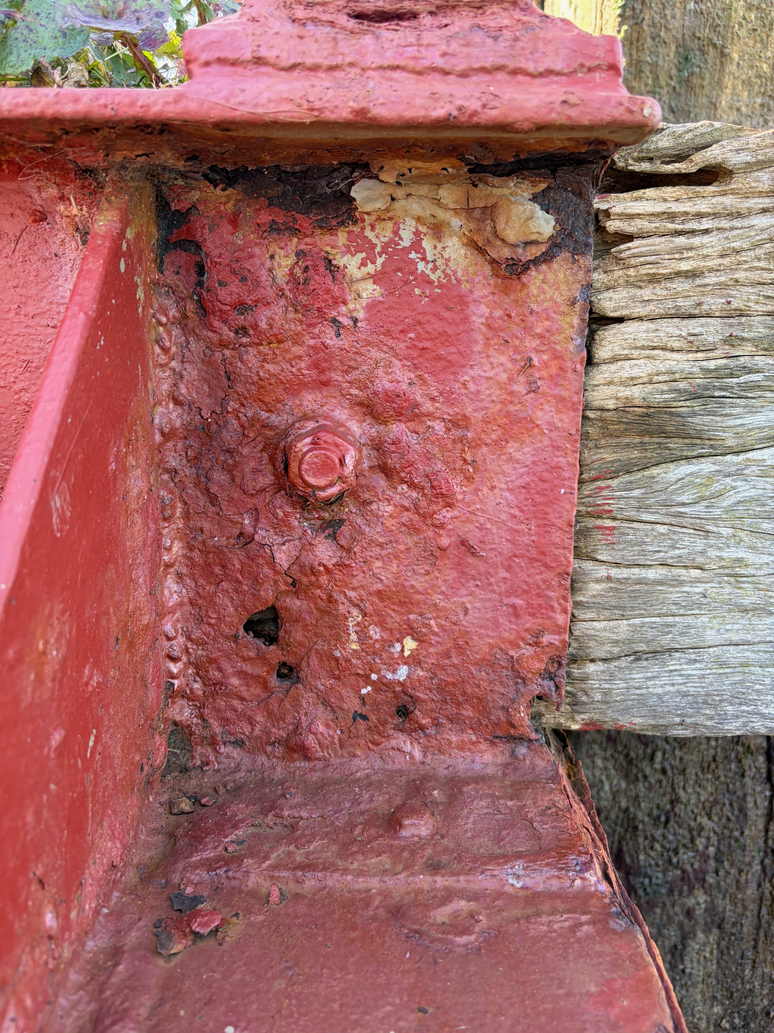 Close-up of a corner of a weathered, red-painted metal structure attached to old, gray wooden planks, showing rust, chipped paint, and corrosion.