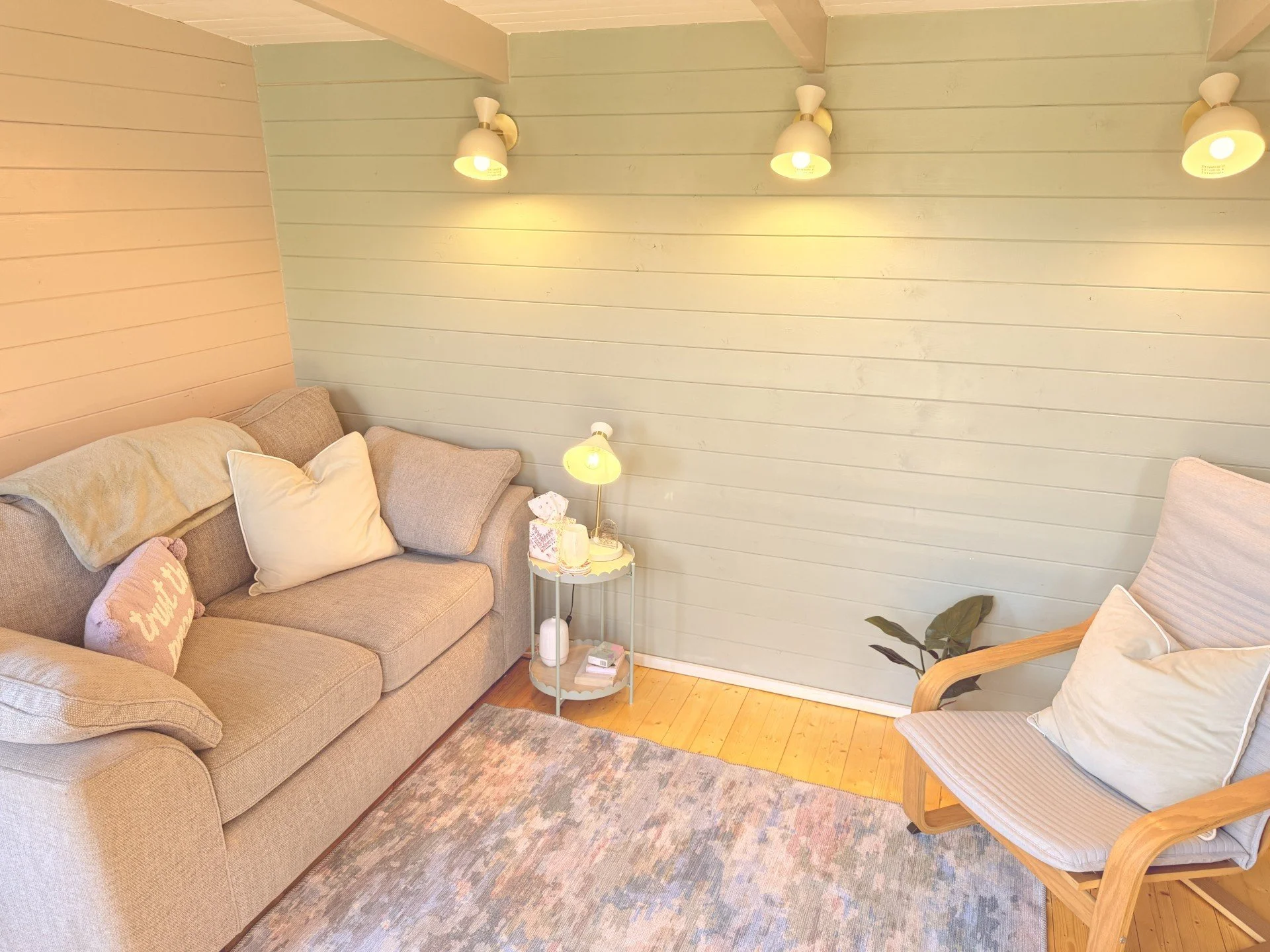 Living room corner with beige sofa, throw pillows, small white side table with lamp and books, wooden chair, and light green paneled walls with wall-mounted lights.