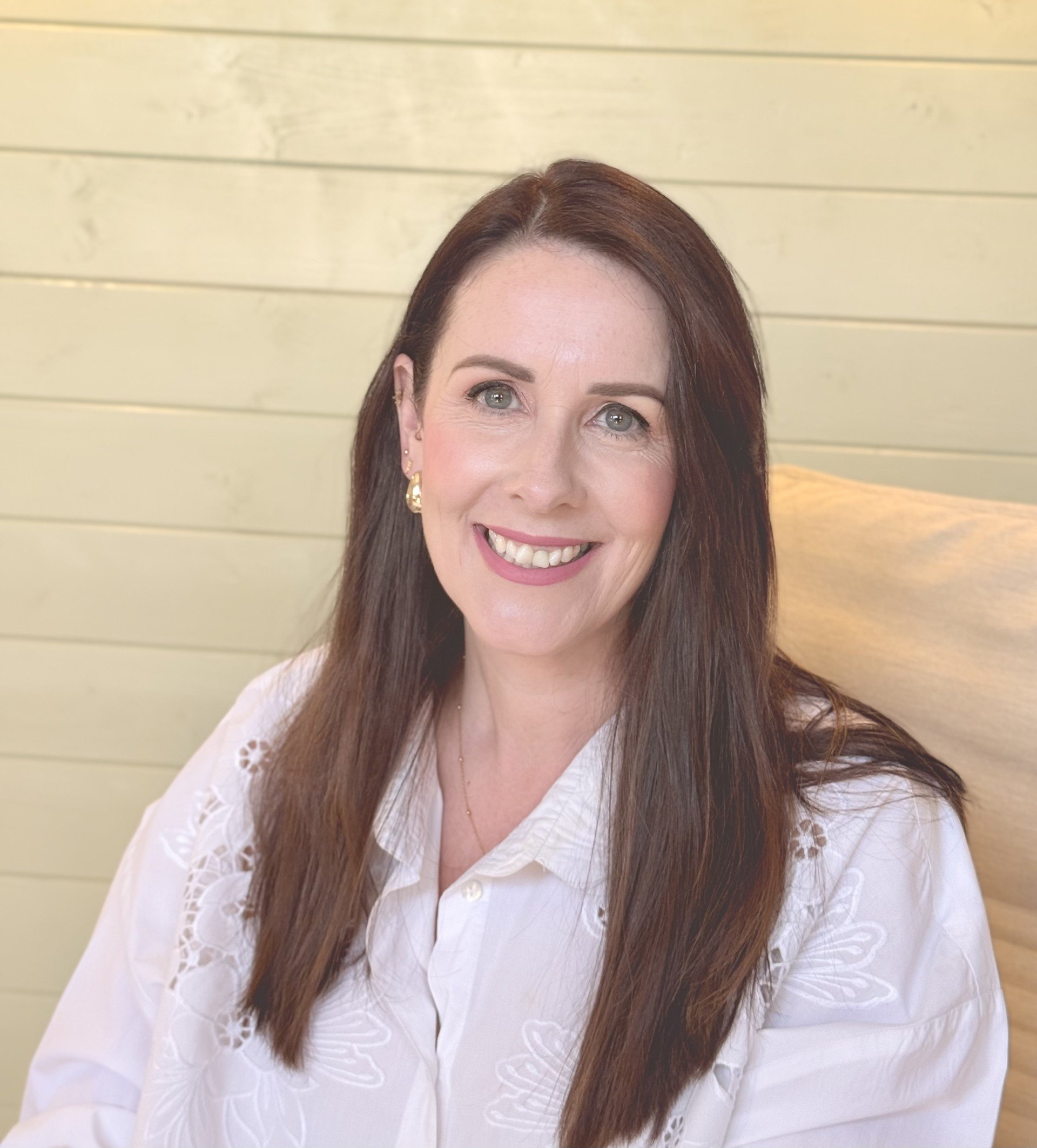 A woman with long brown hair, light skin, and blue eyes smiling, wearing a white blouse with embroidery, seated in front of a light yellow shiplap wall.