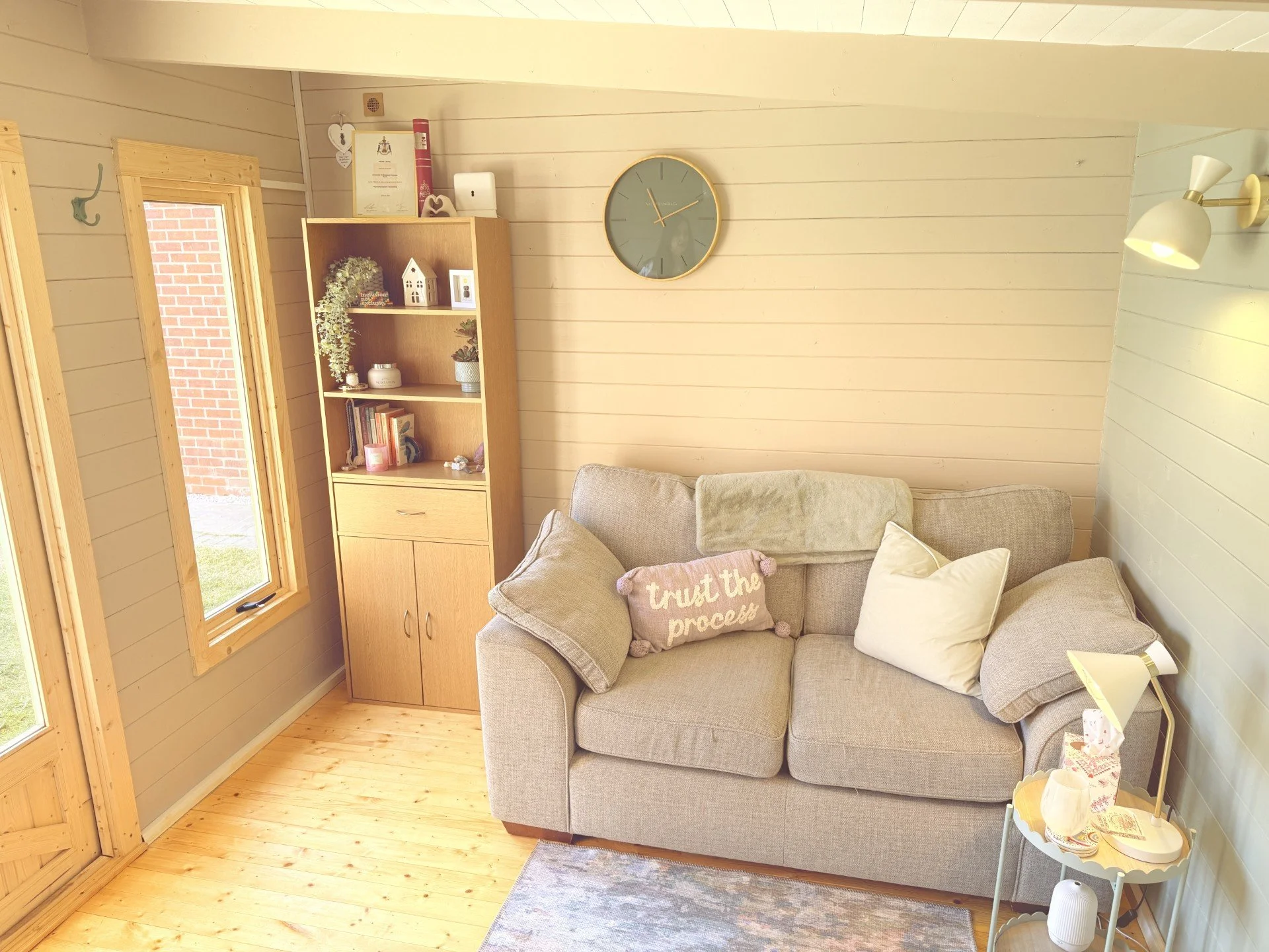 Therapy space at Natalie Clowes Counselling light-colored wooden walls and floor, a gray sofa with beige and pink pillows, a wall clock, a wooden cabinet with decorative items, a window with a brick exterior view, and a floor lamp.