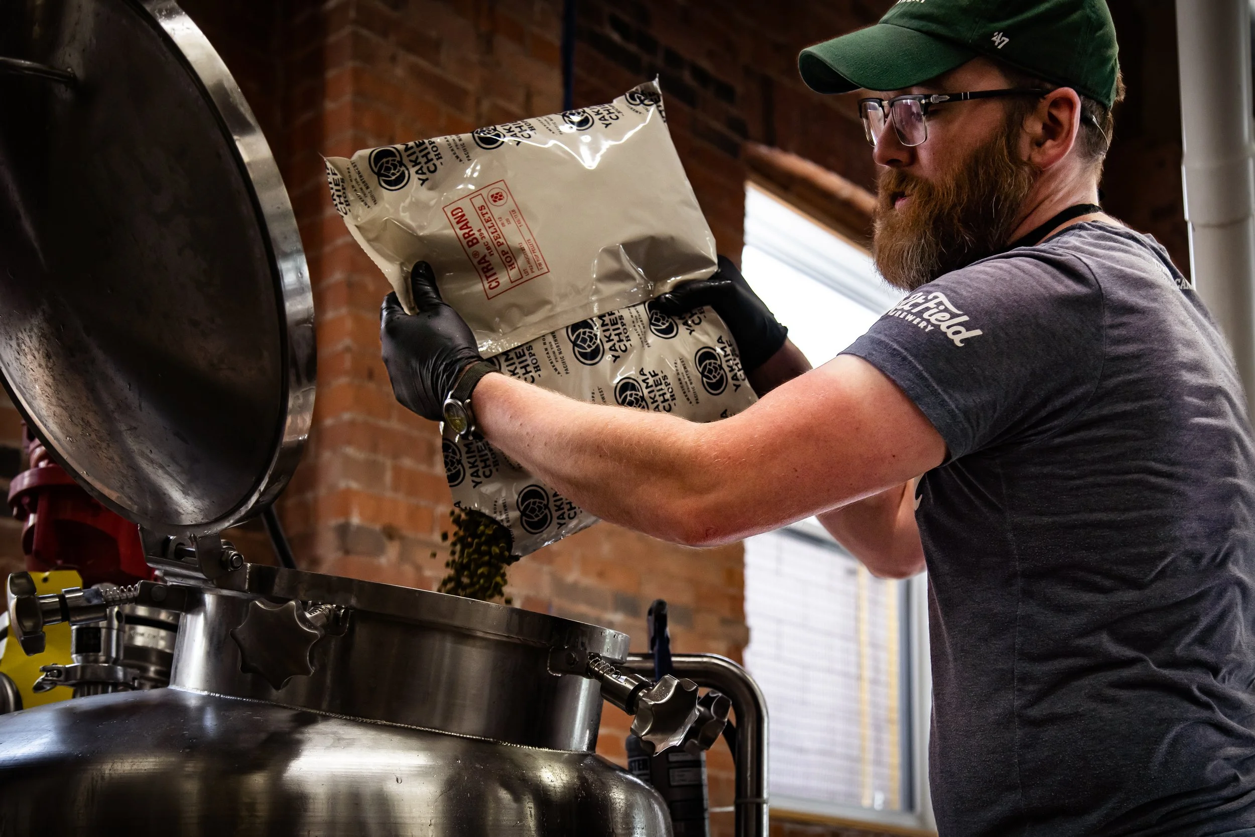 A craft brewer with a beard and glasses pouring a bag of hops into a large vat in a brewery.