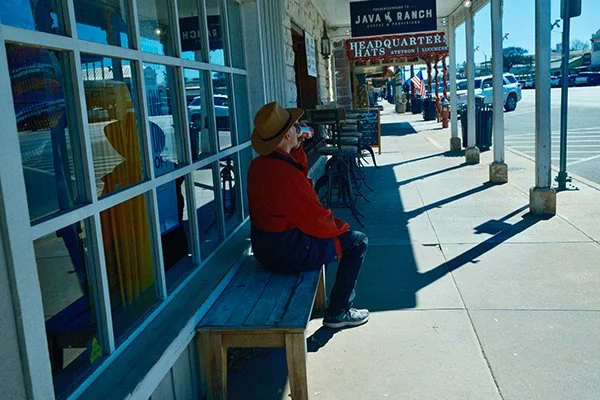Person in a red jacket and hat sitting on a bench outside a shop with large glass windows, reflecting the street scene, with storefront signs and parked cars in the background.