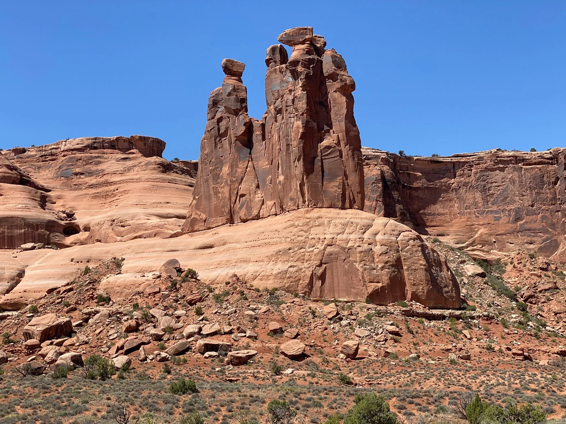Reddish-brown rock formations with tall, thin towers and boulders, under a clear blue sky, sparse desert vegetation at the base.