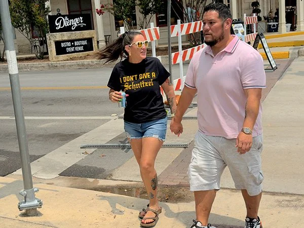 A woman and a man walking hand in hand on a sidewalk in an urban area. The woman is wearing a black T-shirt with yellow lettering, denim shorts, and flip-flops, and has sunglasses and a tattoo on her leg. The man is wearing a light pink polo shirt, g