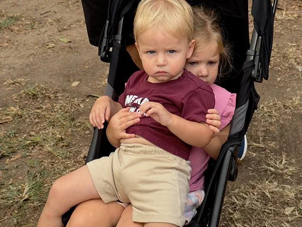 Two young children sitting in a stroller outdoors, with the boy looking directly at the camera and the girl hugging him from behind.