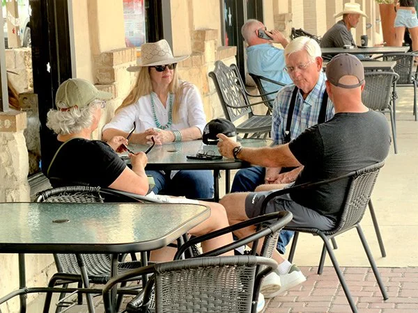 Four older adults sitting outside at a table, two women and two men, engaging in conversation. One man is holding a black hat and the woman in white is wearing a wide-brim hat and sunglasses.