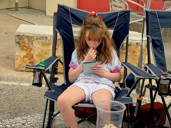 A young girl with long brown hair sitting in a portable camping chair outdoors, looking at her phone with her thumb near her lips. She is wearing a colorful shirt and white shorts. There is a plastic cup in the foreground and some chairs and a stone 