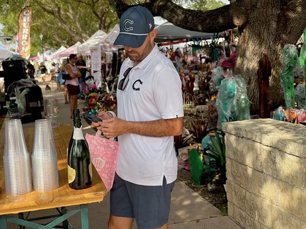 Man in white polo shirt, navy cap, and sunglasses looking at his phone at an outdoor market or festival, with plastic cups and a bottle of champagne on the table beside him, and vendor tents and people in the background.