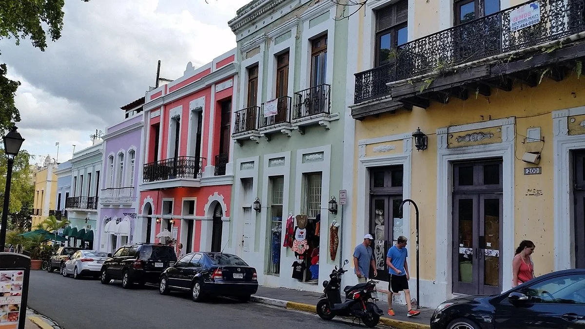 Colorful row of historic buildings on a city street with parked cars and pedestrians, featuring pastel facades and wrought-iron balconies.