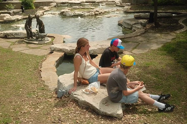 Three children sitting on a large rock near a pond in a park, with two children in the background playing by the water.