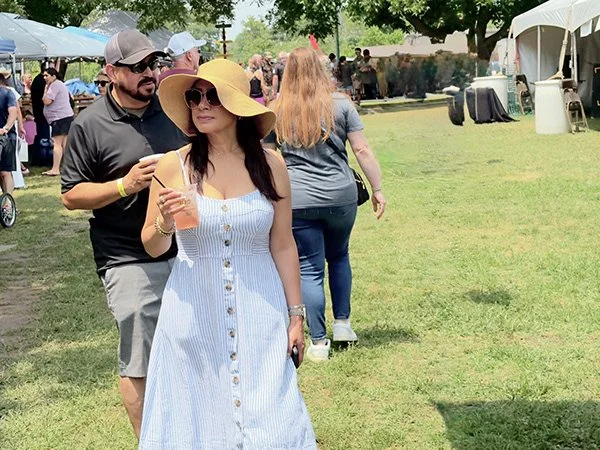 Woman wearing a large sun hat and sunglasses holding a drink walking through an outdoor event with tents and people in the background.