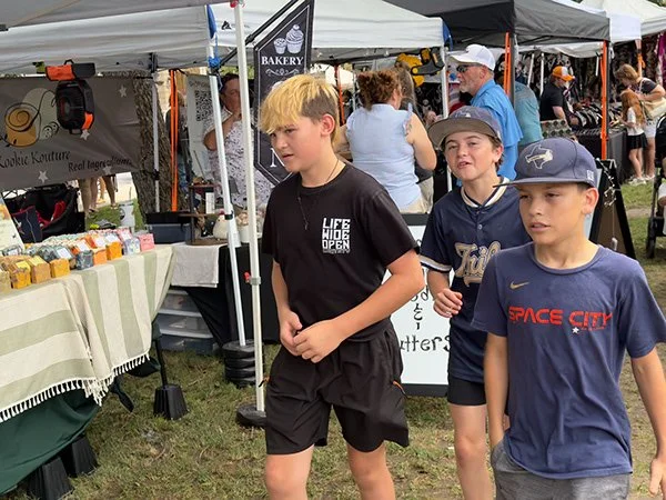 Three young boys walking at an outdoor fair or market with tents and vendor booths behind them.