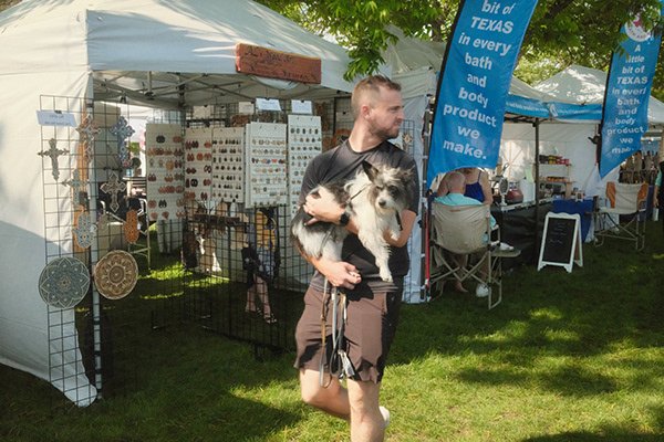 A man is holding a dog at an outdoor market or fair, with tents and display tables in the background.
