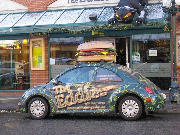 A Volkswagen Beetle car parked outside a burger restaurant called The Eddie Burger Bar, decorated with a giant hamburger on the roof and a large inflatable black dragon hanging above the entrance.