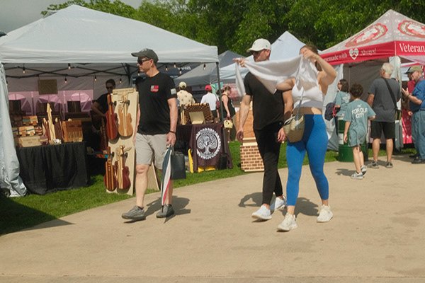 People walking past craft booths at an outdoor market, with tents, guitars, and other items displayed for sale.