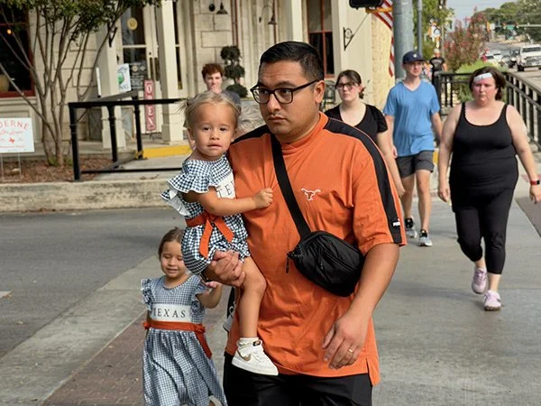Man carrying a young girl dressed in a checkered dress with a blue and white pattern while walking on a city sidewalk, with other pedestrians in the background.