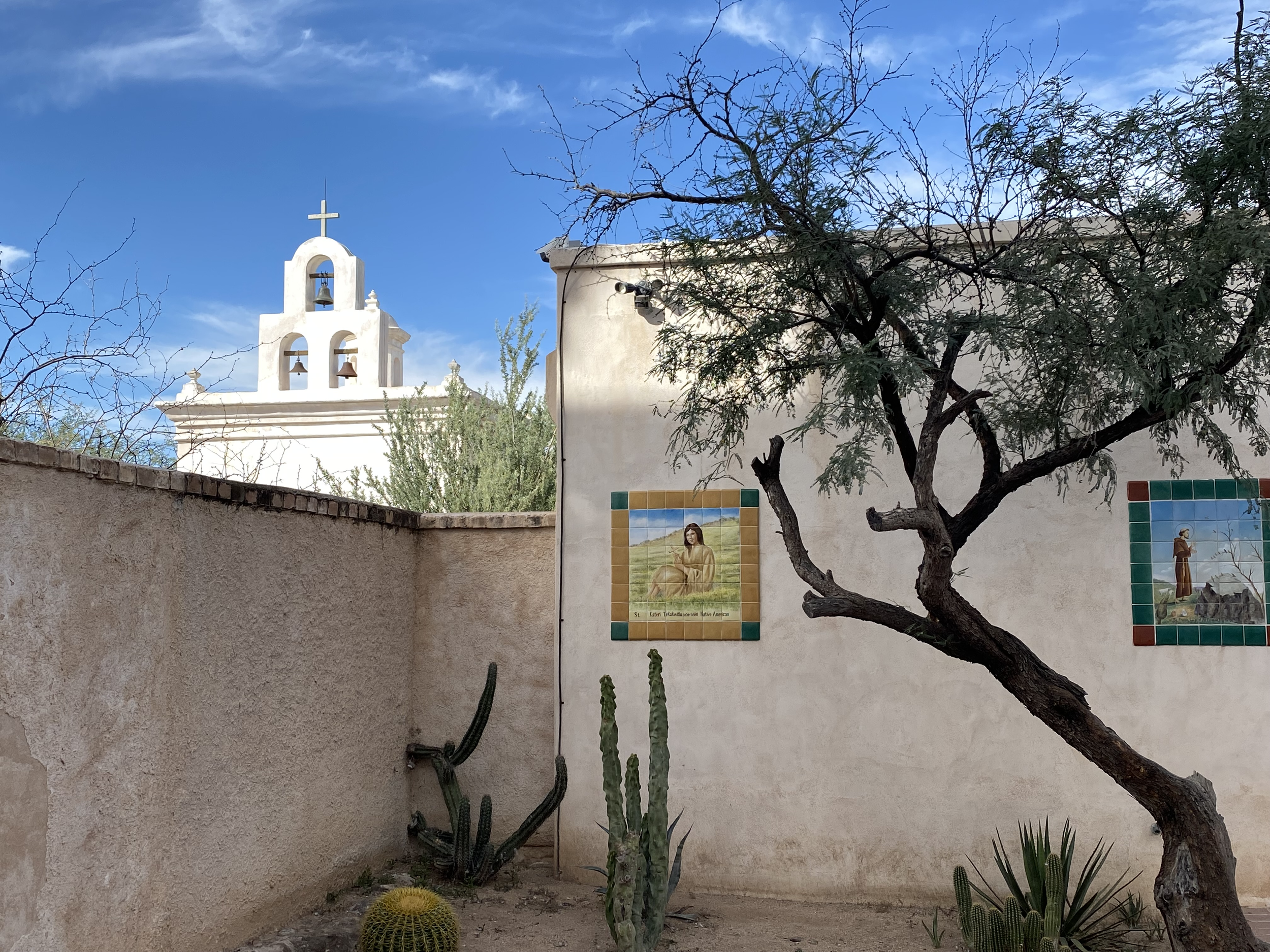 A white church with a bell tower and cross on top, seen behind a beige wall with colorful tiles, trees, cacti, and desert plants in front, under a partly cloudy blue sky.