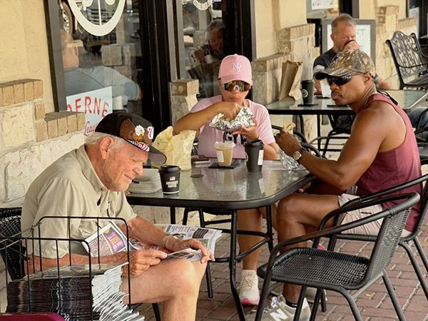 Three people sitting at an outdoor cafe table with coffee cups and food, two men and one girl wearing sunglasses, a pink cap, and a headband.