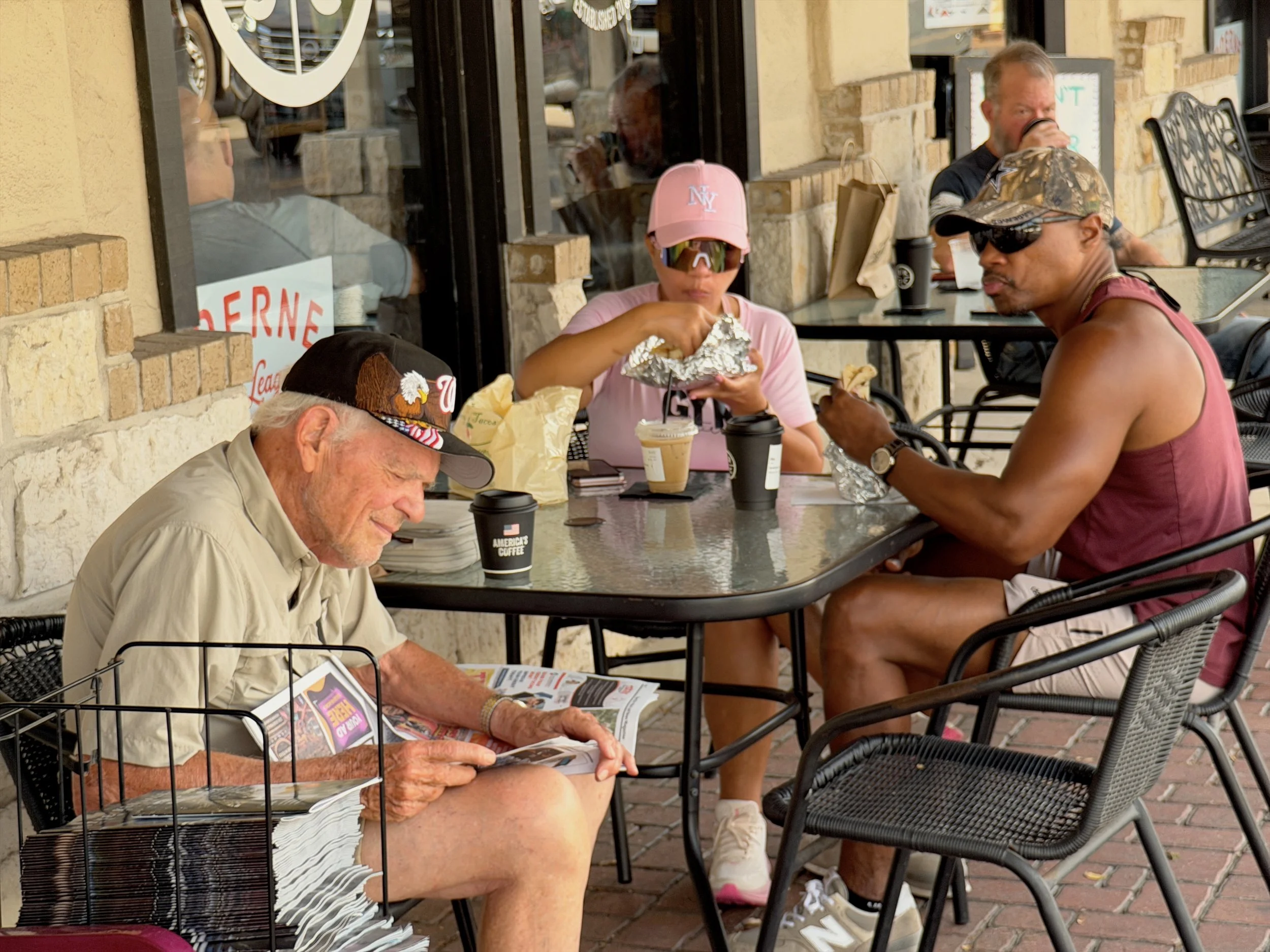 People sitting at an outdoor café, eating and drinking, with a window reflecting the street and a sign that reads 'OPEN' in the background.