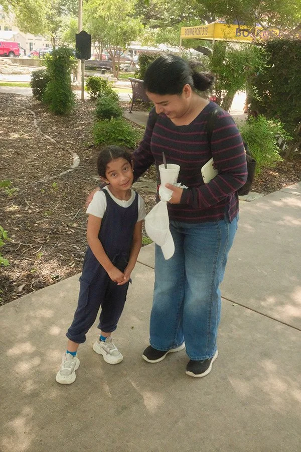 A woman and a young girl standing outdoors on a sidewalk in a park-like area, smiling at each other. The woman is holding a drink and a napkin, while the girl is wearing a navy blue outfit with white sneakers. Trees and a street with vehicles are vis
