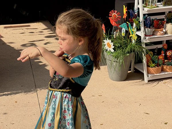 A young girl wearing a dress with floral and striped patterns, holding a string with a tiny object at the end, standing outside near colorful decorative flowers on shelves.