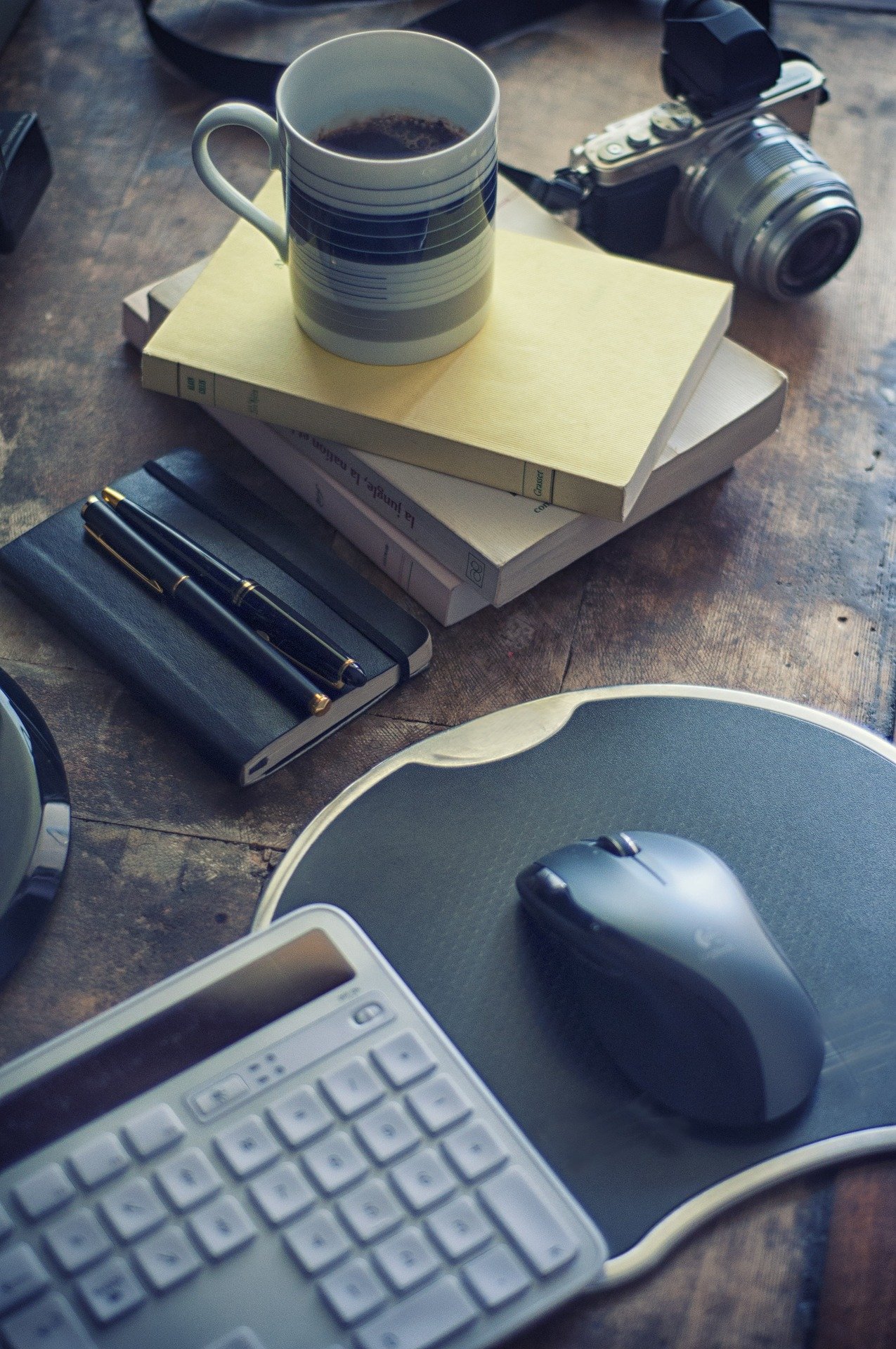 Desk surface with a coffee mug, a camera, a closed notebook, a book, a pen, a wireless mouse, a printer, and a keyboard.