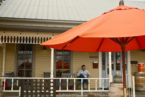A person sitting on the porch of a yellow building under a large orange patio umbrella.