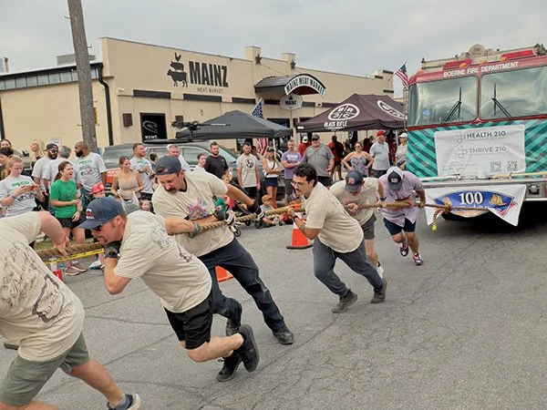 People participating in a tug-of-war competition in front of a crowd and a fire truck, with a building featuring a sign that says 'Mainz' in the background.