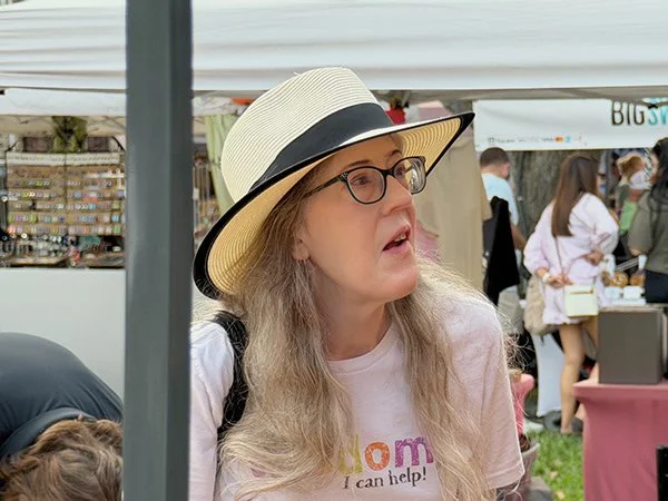A woman wearing a wide-brimmed hat, glasses, and a light pink t-shirt, appears to be at an outdoor event or market, speaking or reacting to something.