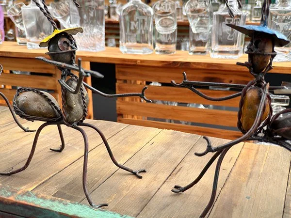 Two metal scarecrow-like figures on a wooden table, one with a yellow hat and the other with a blue hat, holding a stick between them.