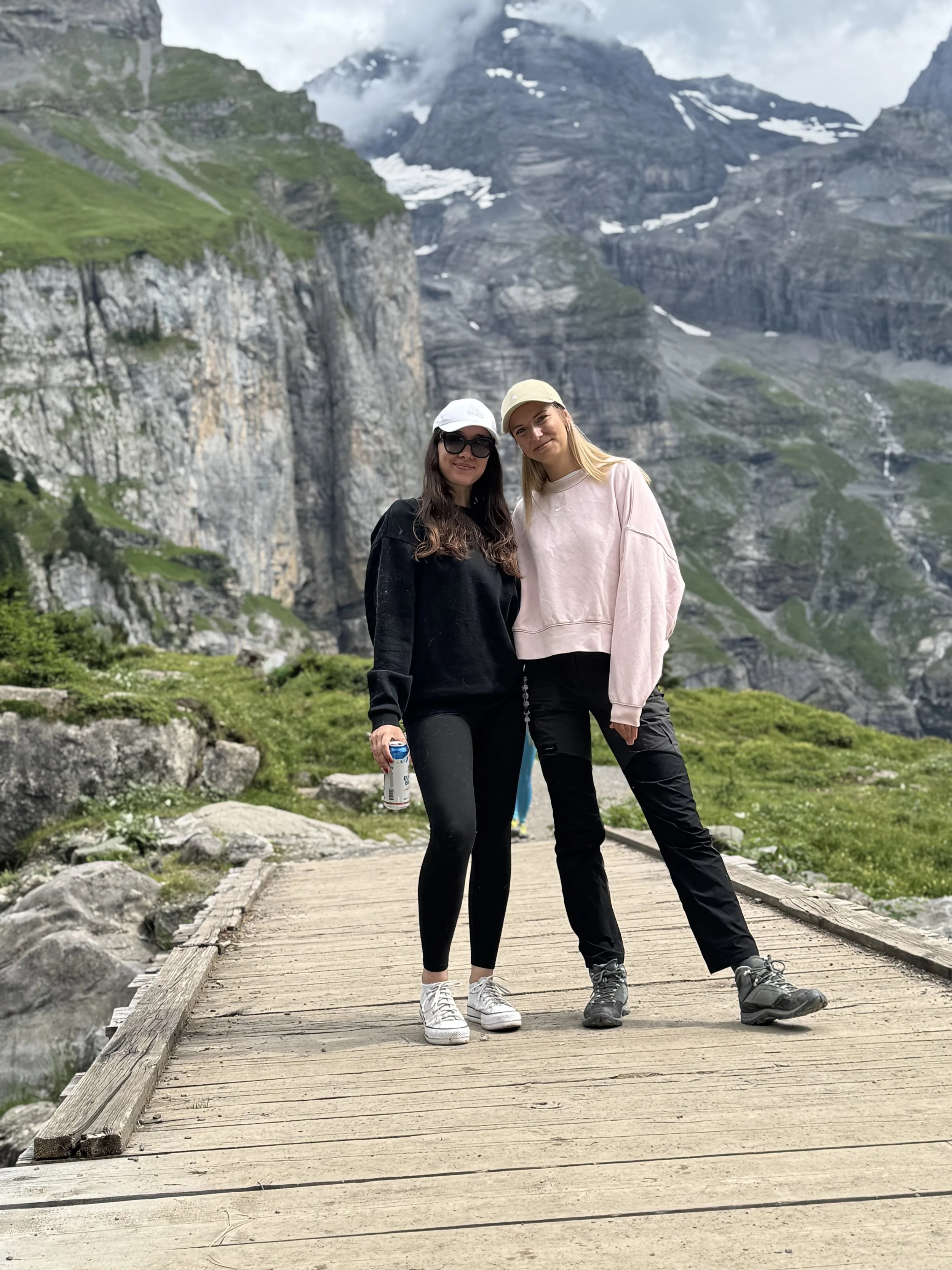 Two women standing on a wooden bridge in a mountainous area with patches of snow, green landscape, and cloudy sky in the background.