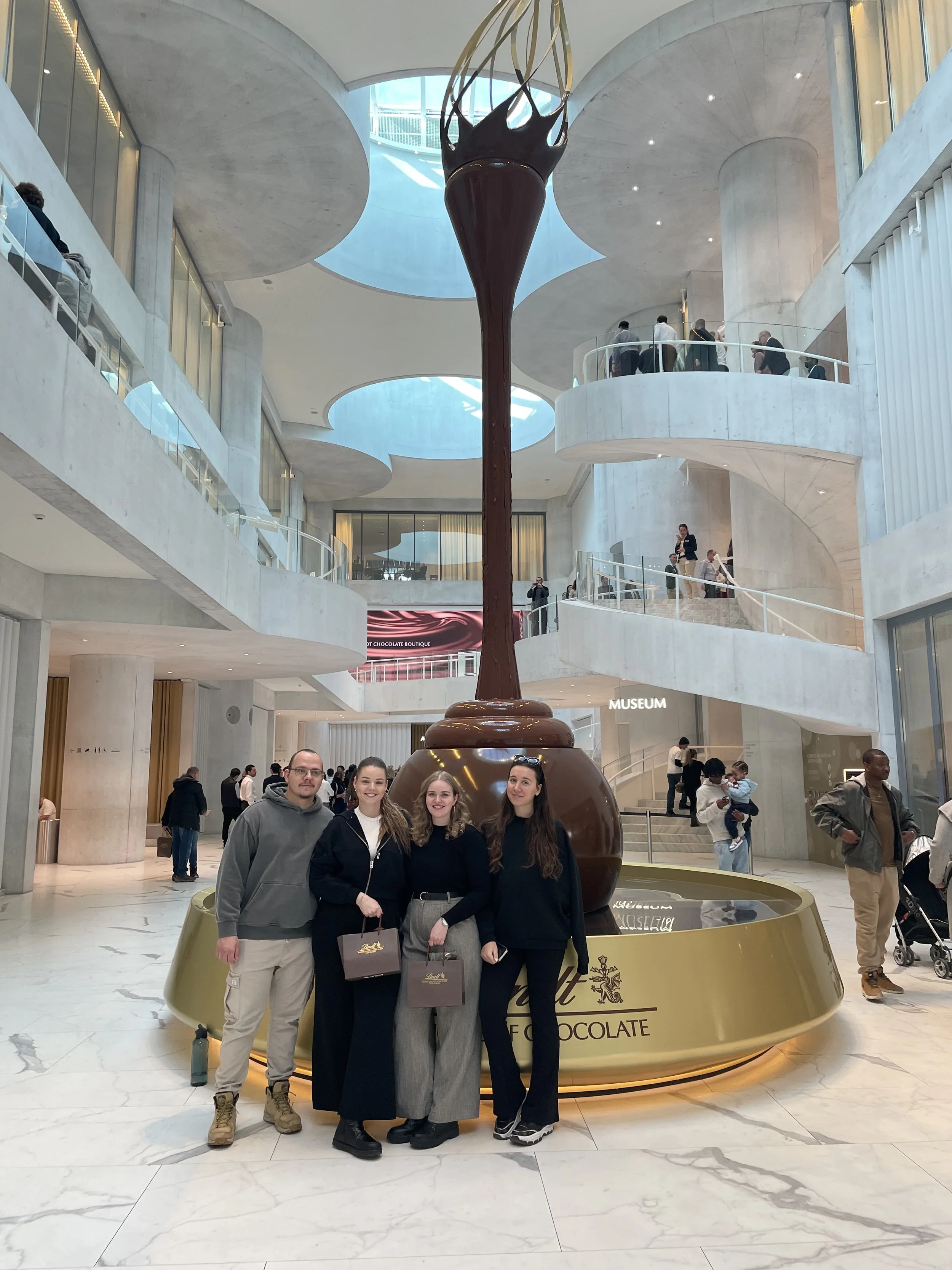 A group of four people standing in front of a large decorative chocolate fountain at an indoor shopping mall. The background includes multiple levels with people walking, modern architectural design, and a sign that says 'MUSEUM.'