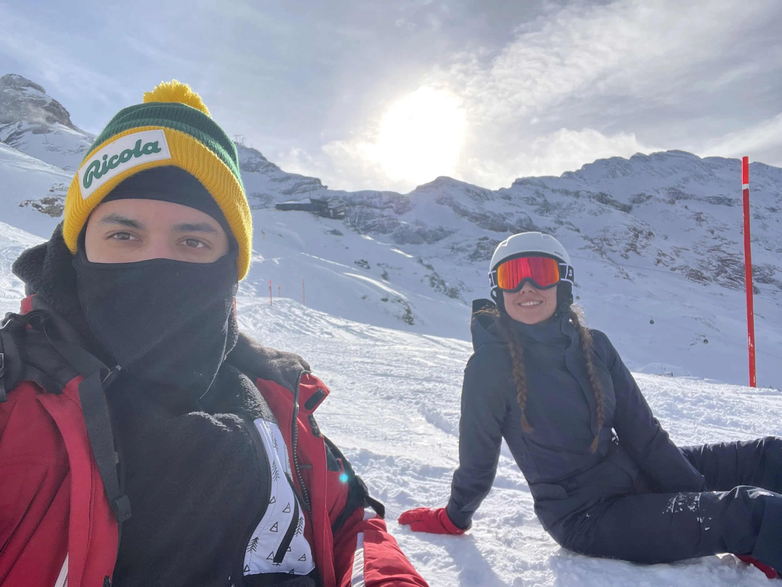 Two people in snow gear on a snowy mountain with ski poles, one wearing a yellow, green, and blue hat with a Ricola logo, and the other with ski goggles and a helmet, smiling with mountains and snow in the background.