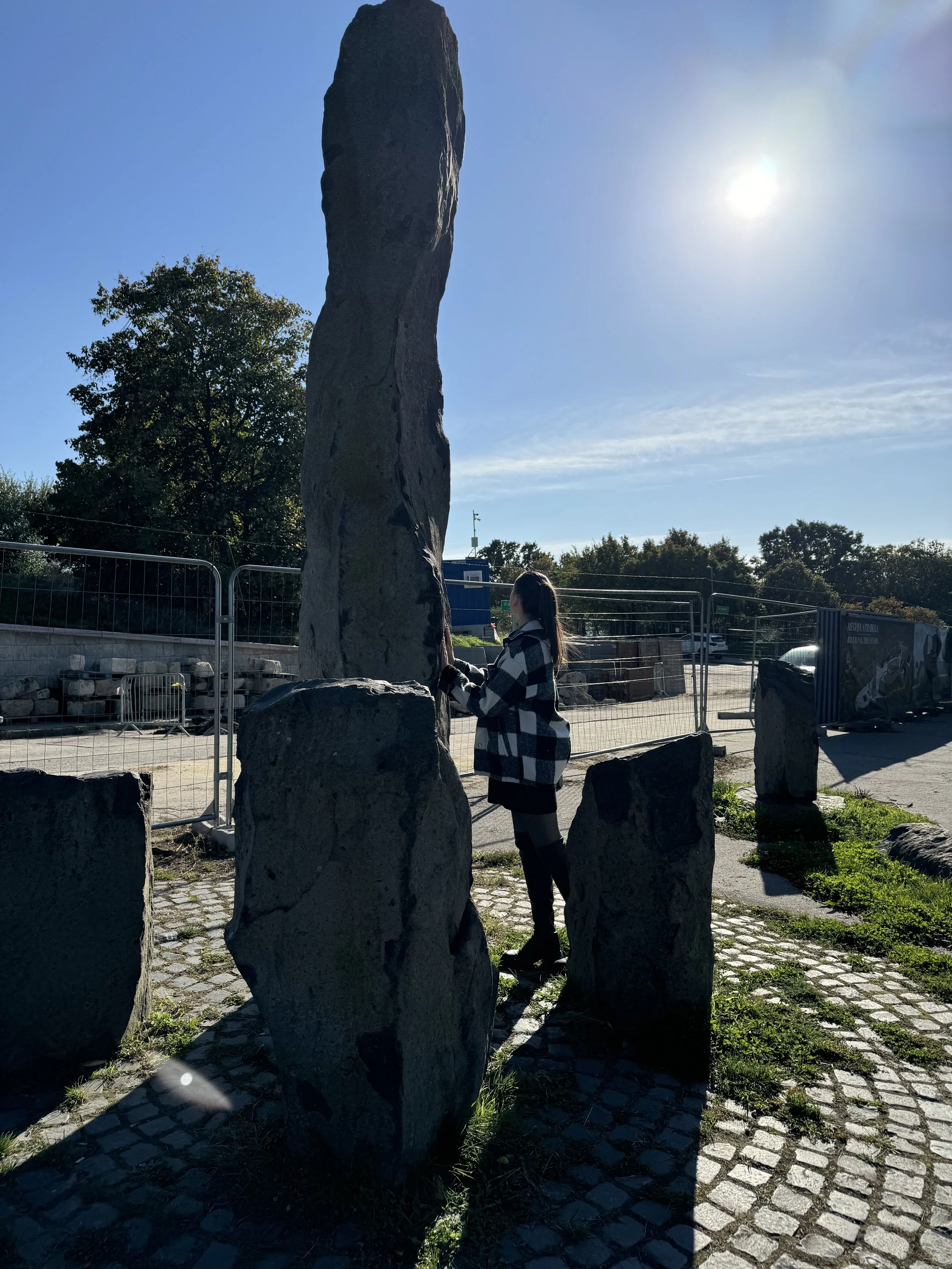 A person in a checkered jacket standing outdoors next to large black stones, with a tall rock formation in the background and the sun shining brightly in the clear sky.