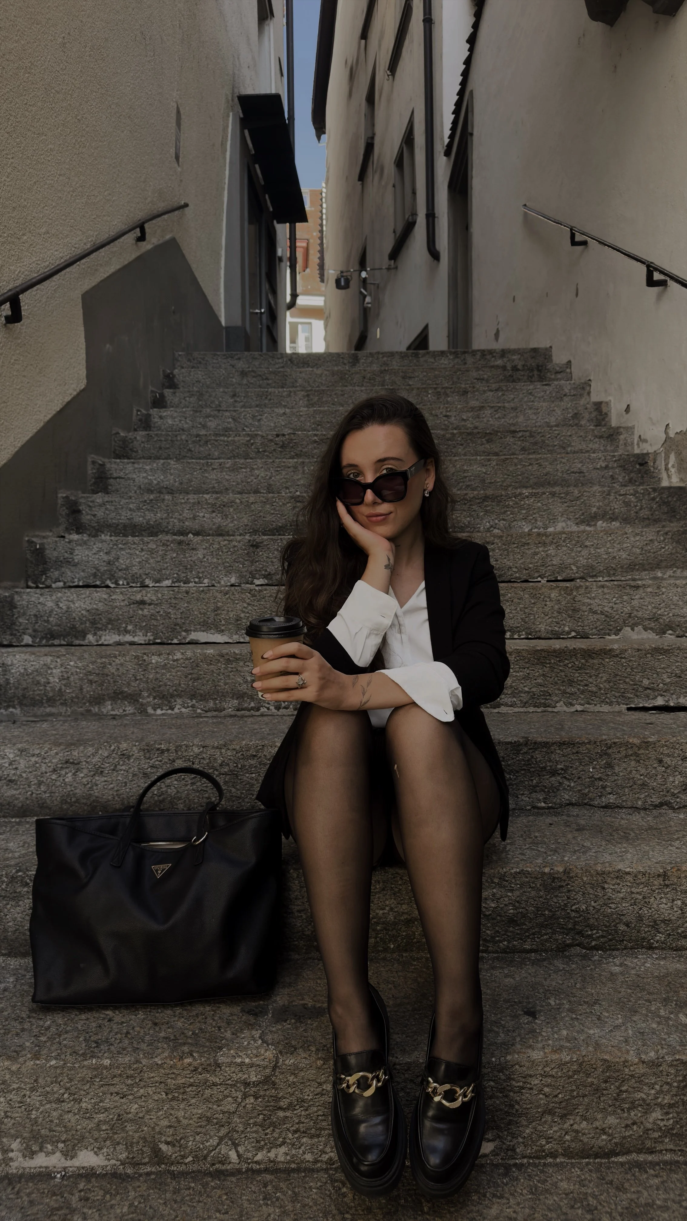 A woman sitting on outdoor stone stairs, wearing black sunglasses, a black blazer, a white shirt, black stockings, and black shoes with gold chain decoration. She holds a coffee cup and has a black handbag beside her.
