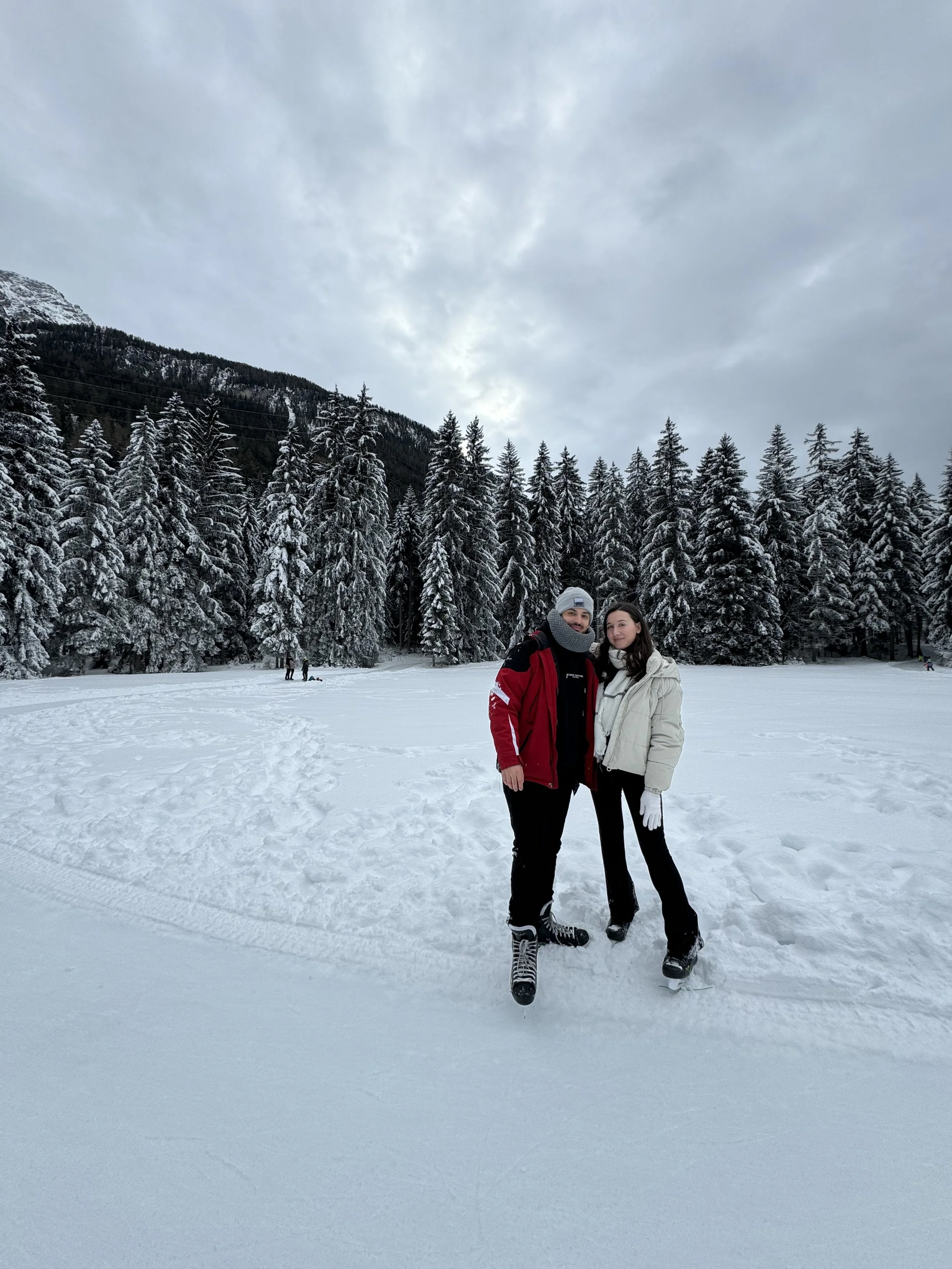A couple dressed in winter clothing standing on snow in a forest with snow-covered trees and a mountain in the background.