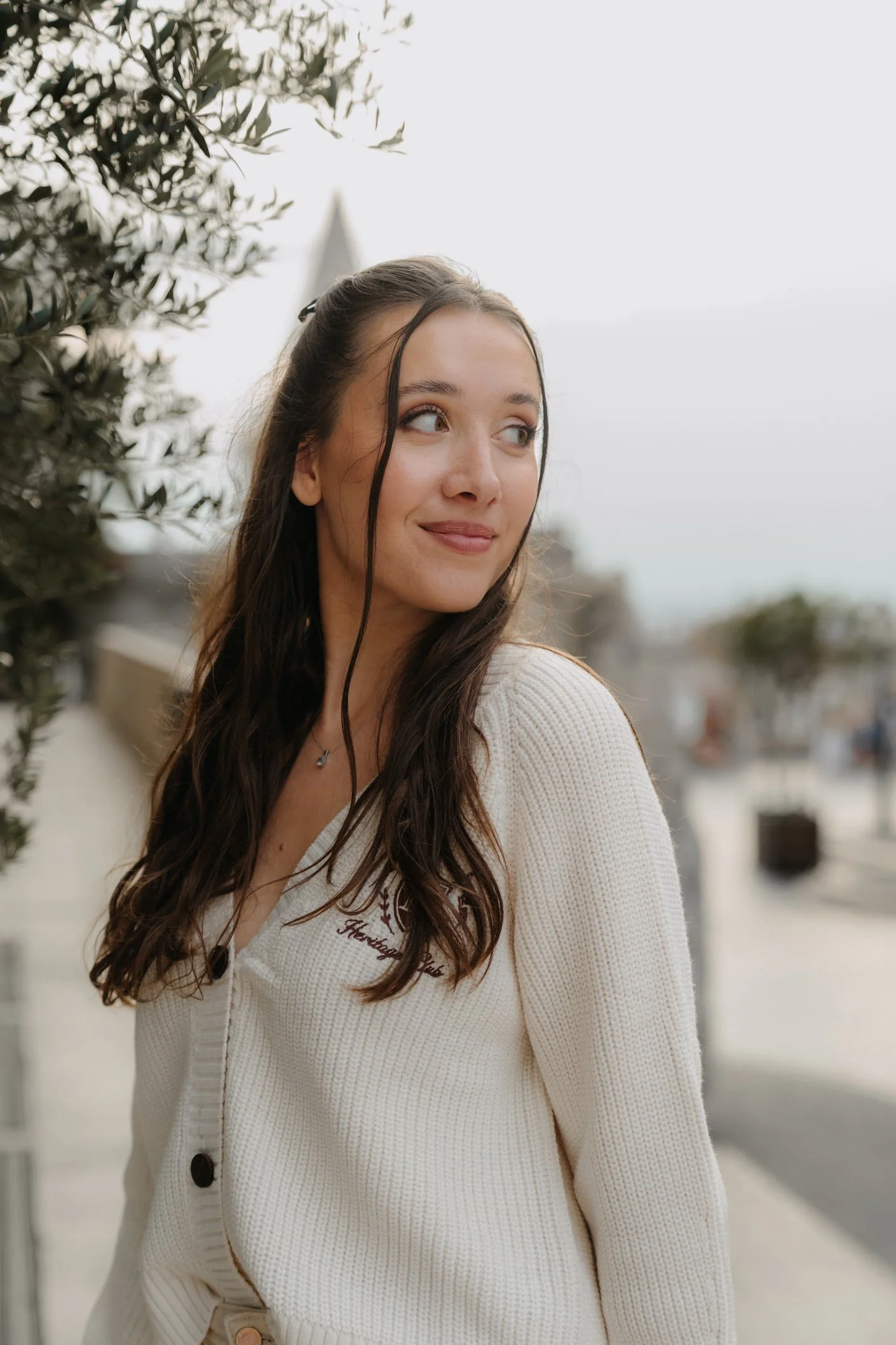 A young woman with long brown hair and light skin is smiling slightly while looking to her right. She is wearing a white sweater with embroidery that says "Huntington Beach." Behind her, there is a blurred cityscape and sky, with some trees and buildings visible.